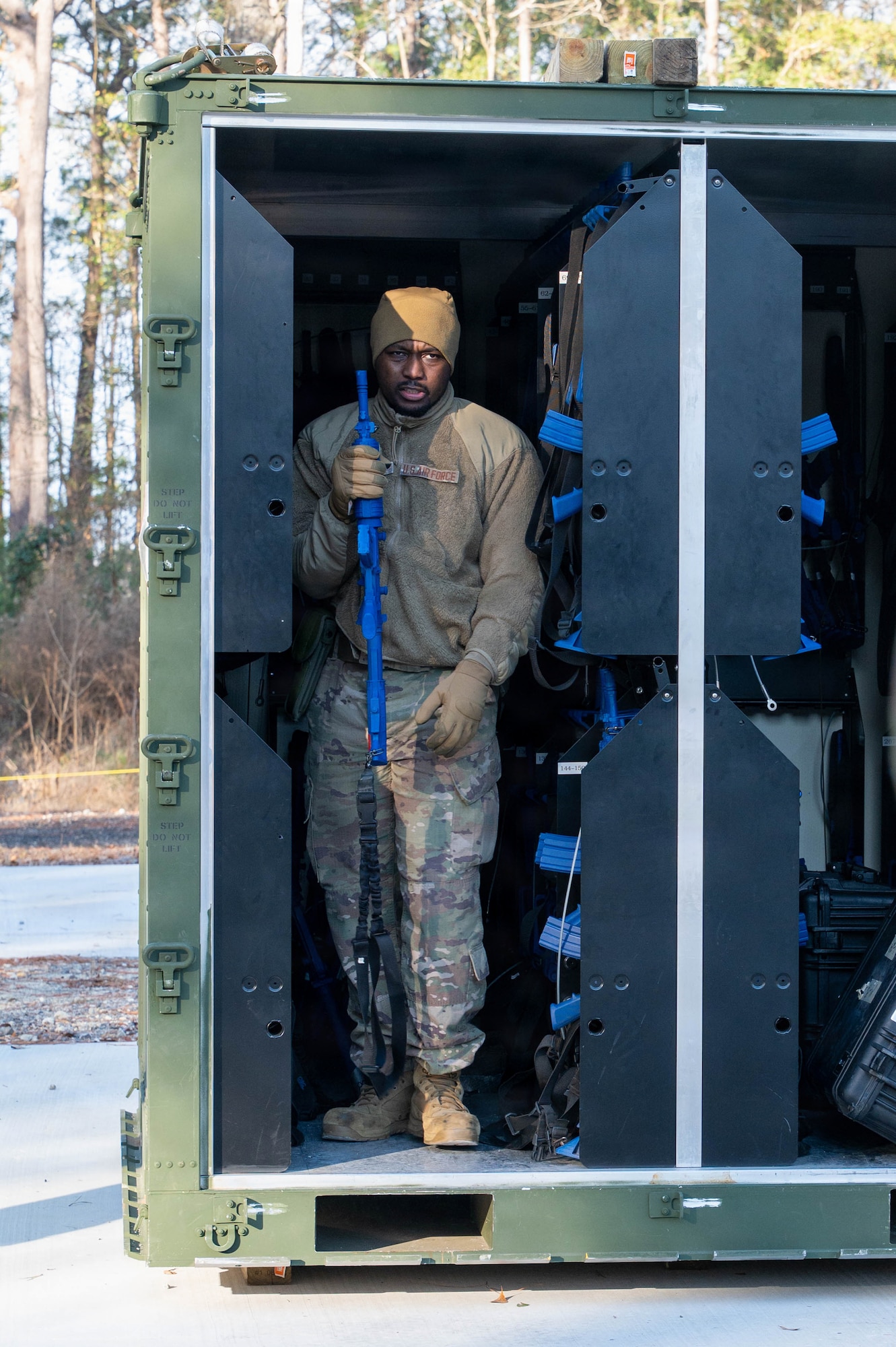 An Airman walks out of a storage container with a blue plastic weapon in hand, the container is lined with storage for blue plastic weapons.