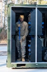 An Airman walks out of a storage container with a blue plastic weapon in hand, the container is lined with storage for blue plastic weapons.