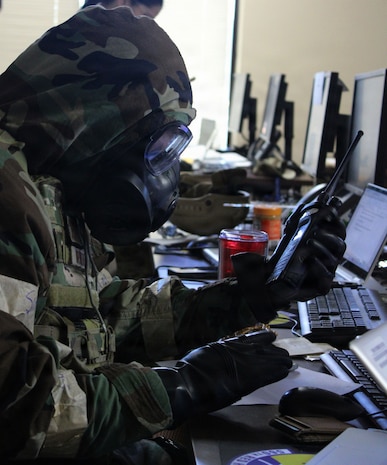 An Airman wearing a gas mask and protective gear holds a walkie talkie in front of his face,