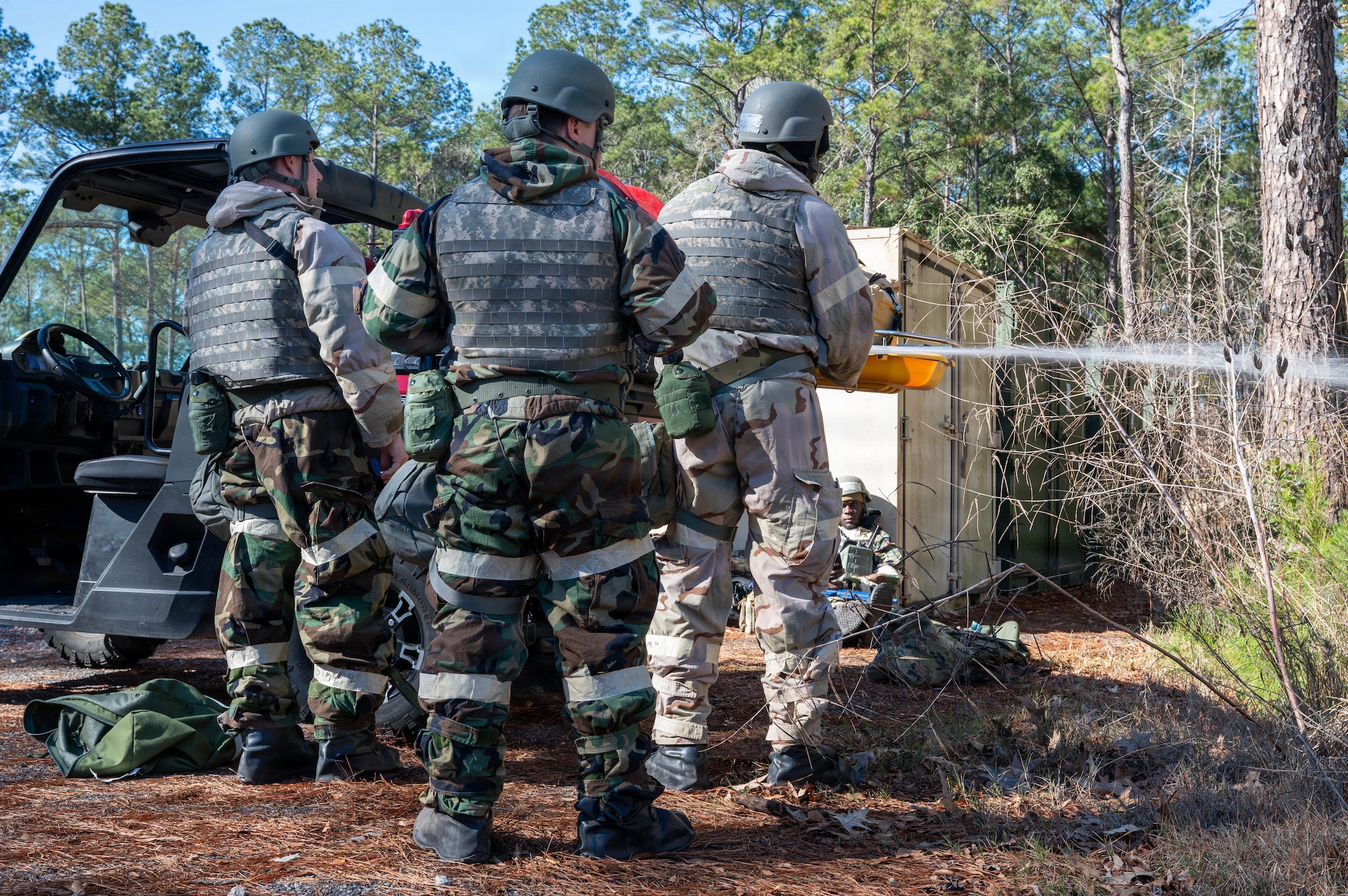 Three Airmen stand in front of a vehicle with their backs to the camera, on the right one of them sprays a hose off to the right.