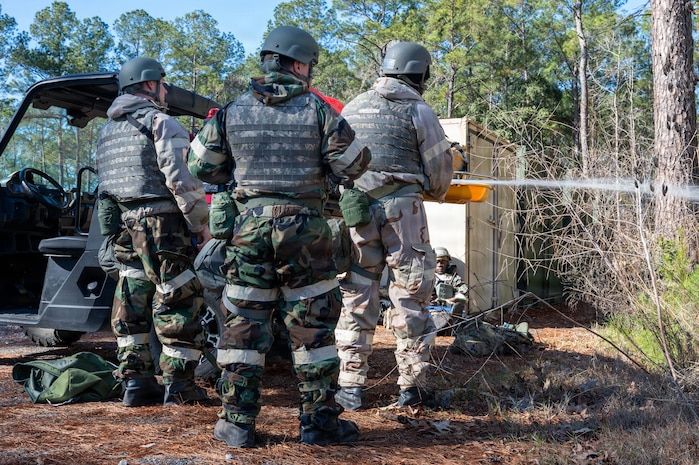 Three Airmen stand in front of a vehicle with their backs to the camera, on the right one of them sprays a hose off to the right.