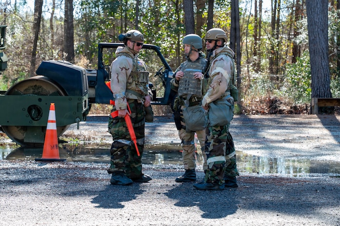 Three Airmen stand in a clearing with equipment in the background.
