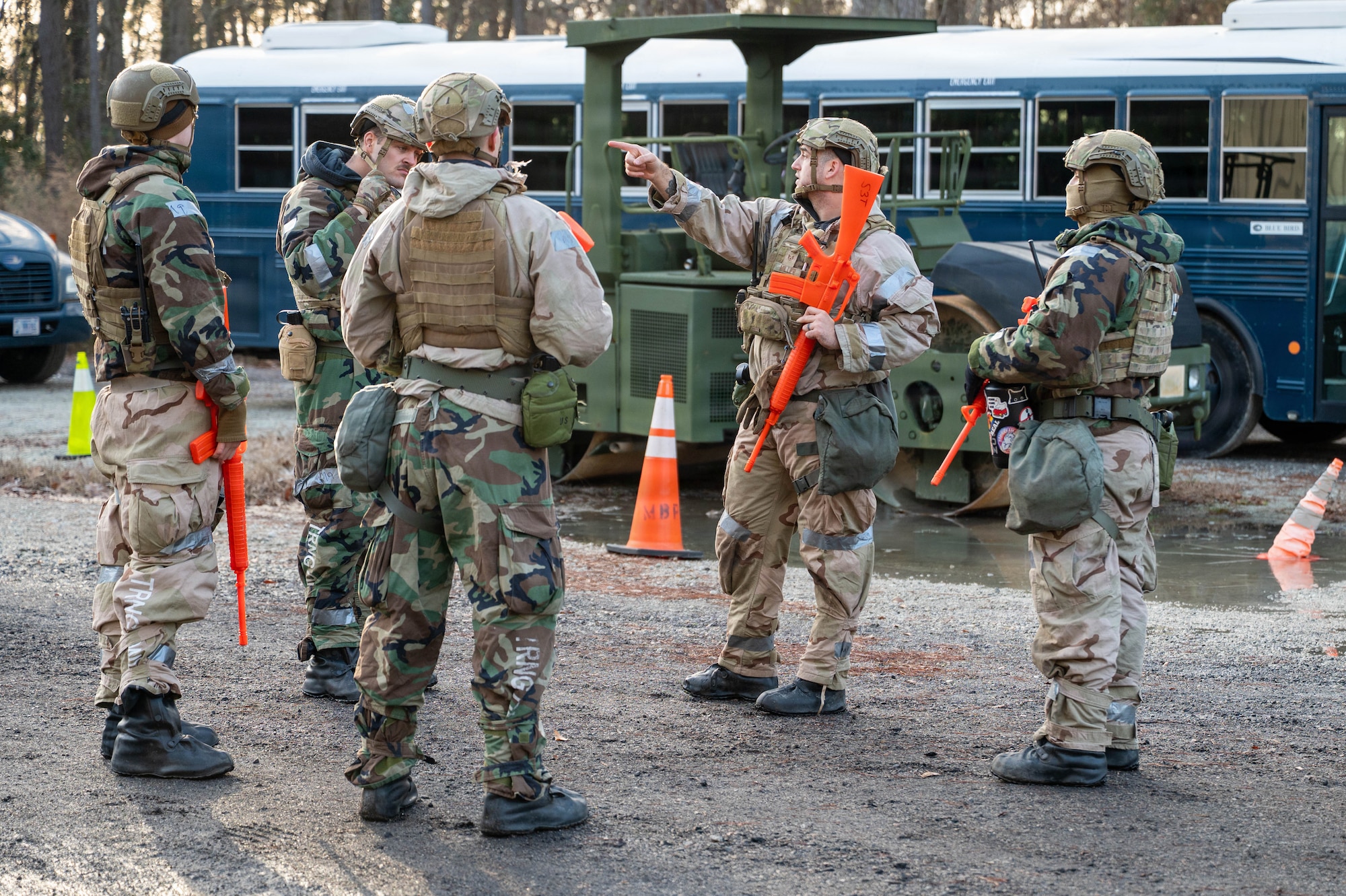 A group of Airmen holding orange plastic weapons stand in a clearing with a blue bus in the background, while one points to the left