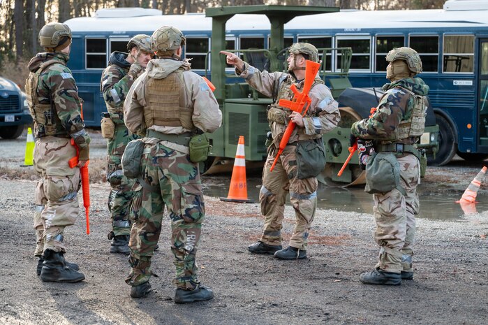 A group of Airmen holding orange plastic weapons stand in a clearing with a blue bus in the background, while one points to the left