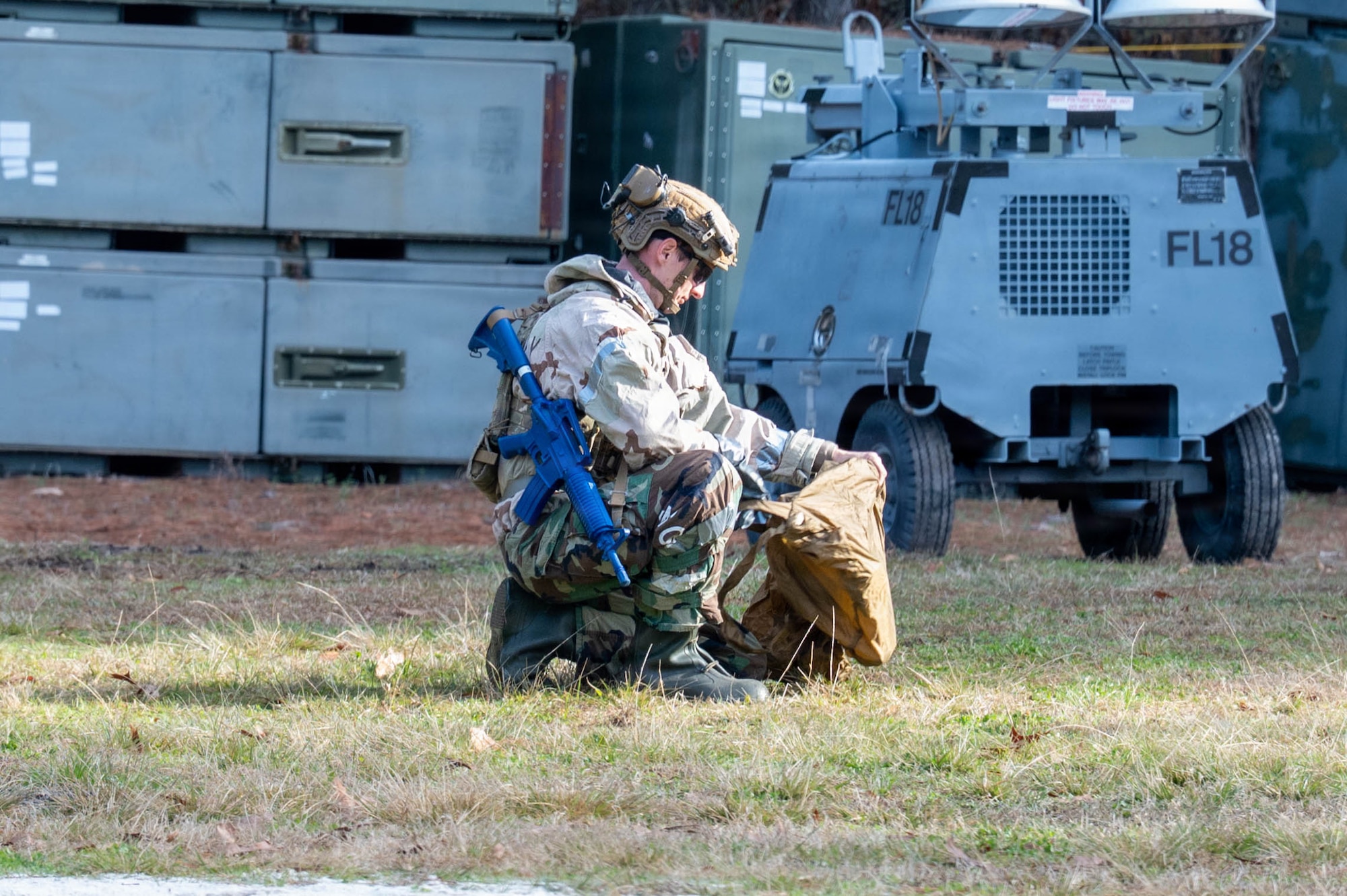 An Airman with a helmet on and a blue plastic weapon around his shoulder kneels on the ground both holding up and putting something into a brown bag