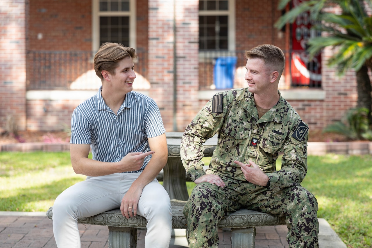 A sailor sits next to a civilian outside on a bench during the day; they are pointing to one another.