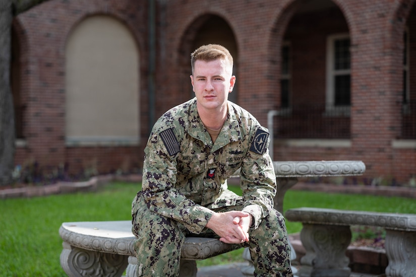 A sailor sits outside on a bench during a sunny day in front of a building.
