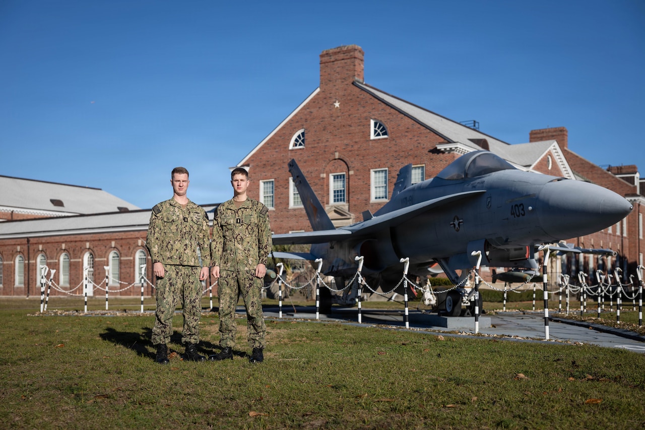 Two men in U.S. Navy uniforms stand side-by-side outside on a clear day next to a parked military aircraft.