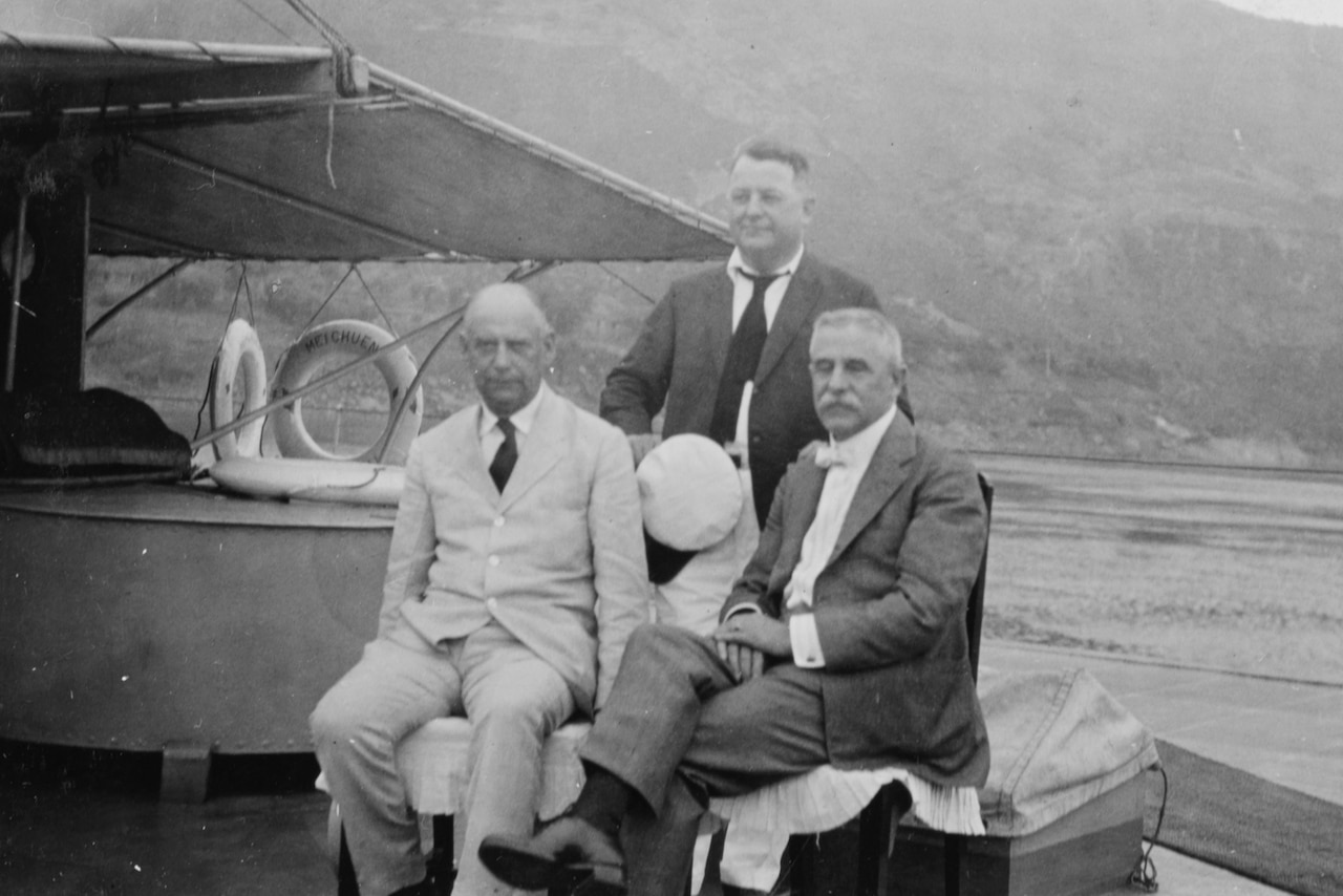Three men in business attire sit on a boat with mountains in the background.