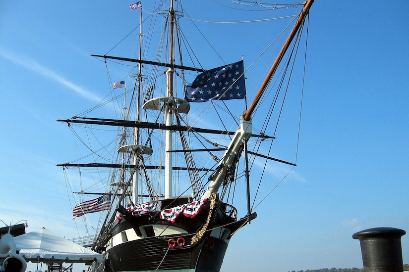 A large military sailing ship flying the American flag and the Union Jack is anchored beside a seawall.