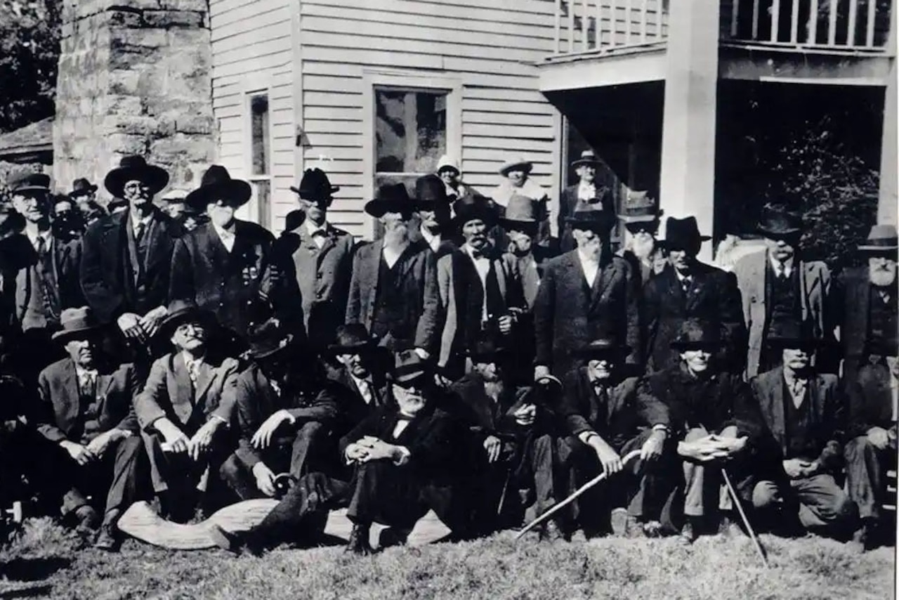 Old men wearing suits and hats stand and sit in two rows as they pose for a photo in front of a building with a stone chimney.