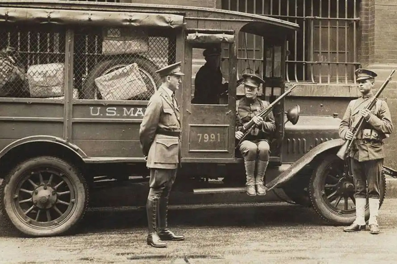 Three Marines in military uniforms guard a U.S. mail truck in front of a large building. Two of the Marines are carrying rifles, while one of them is sitting in the truck. The other Marine is standing next to the truck with his arms behind his back.