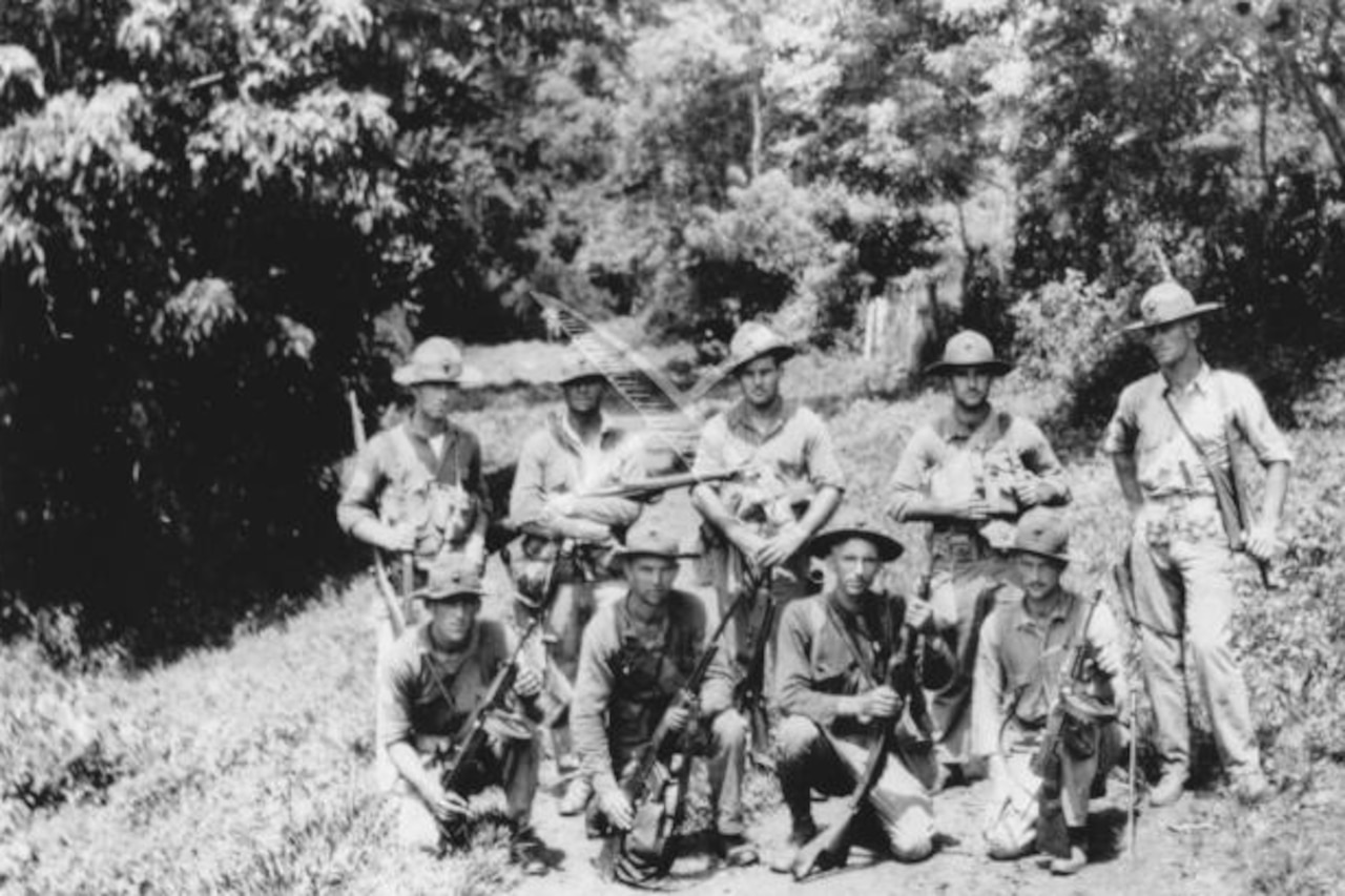 Marines in battle gear with a variety of guns stand and kneel in two rows as they pose for a photo on a dirt road in the jungle.