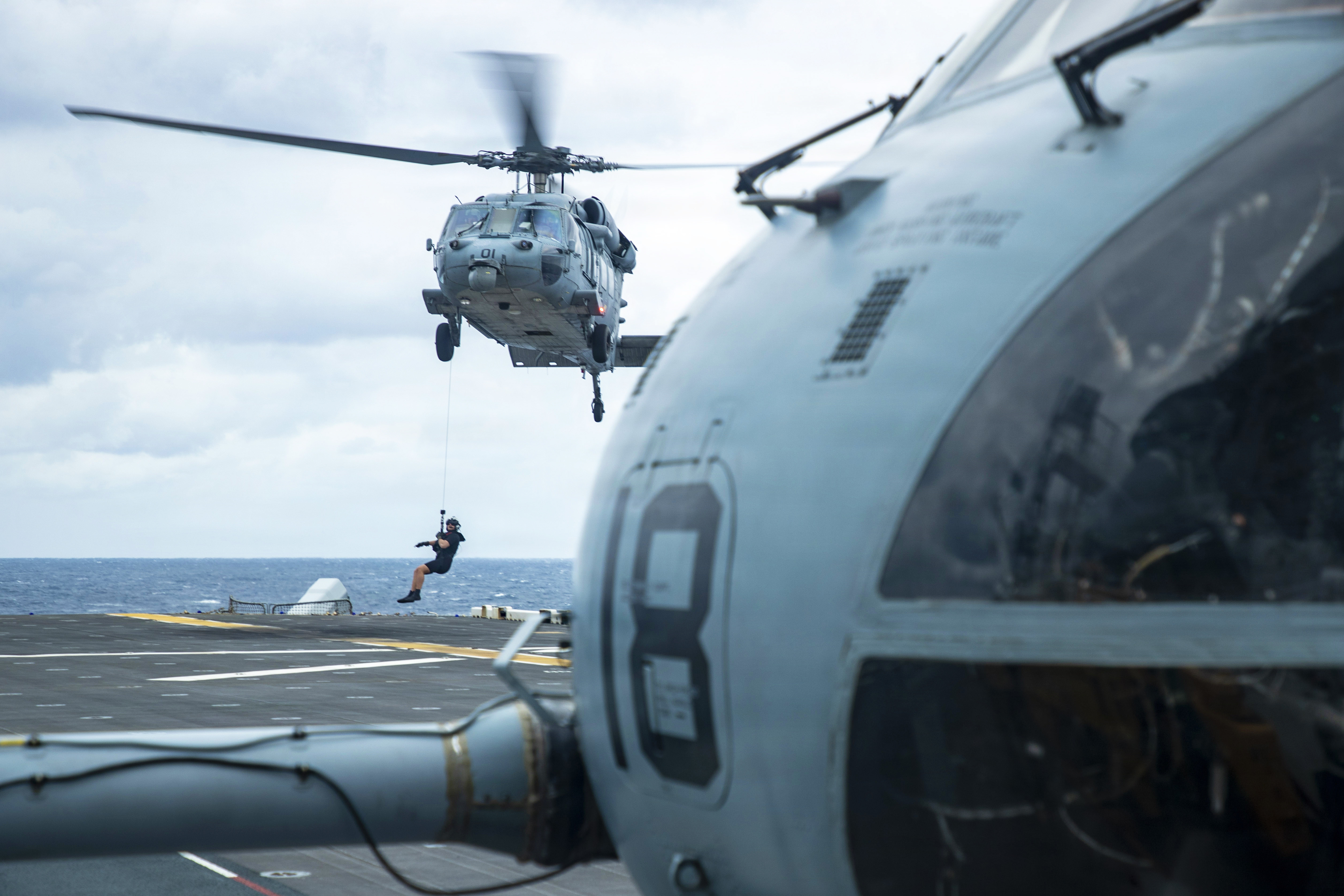 A sailor rappels from a helicopter that hovers over the deck of a ship with part of another helicopter visible in the right foreground.