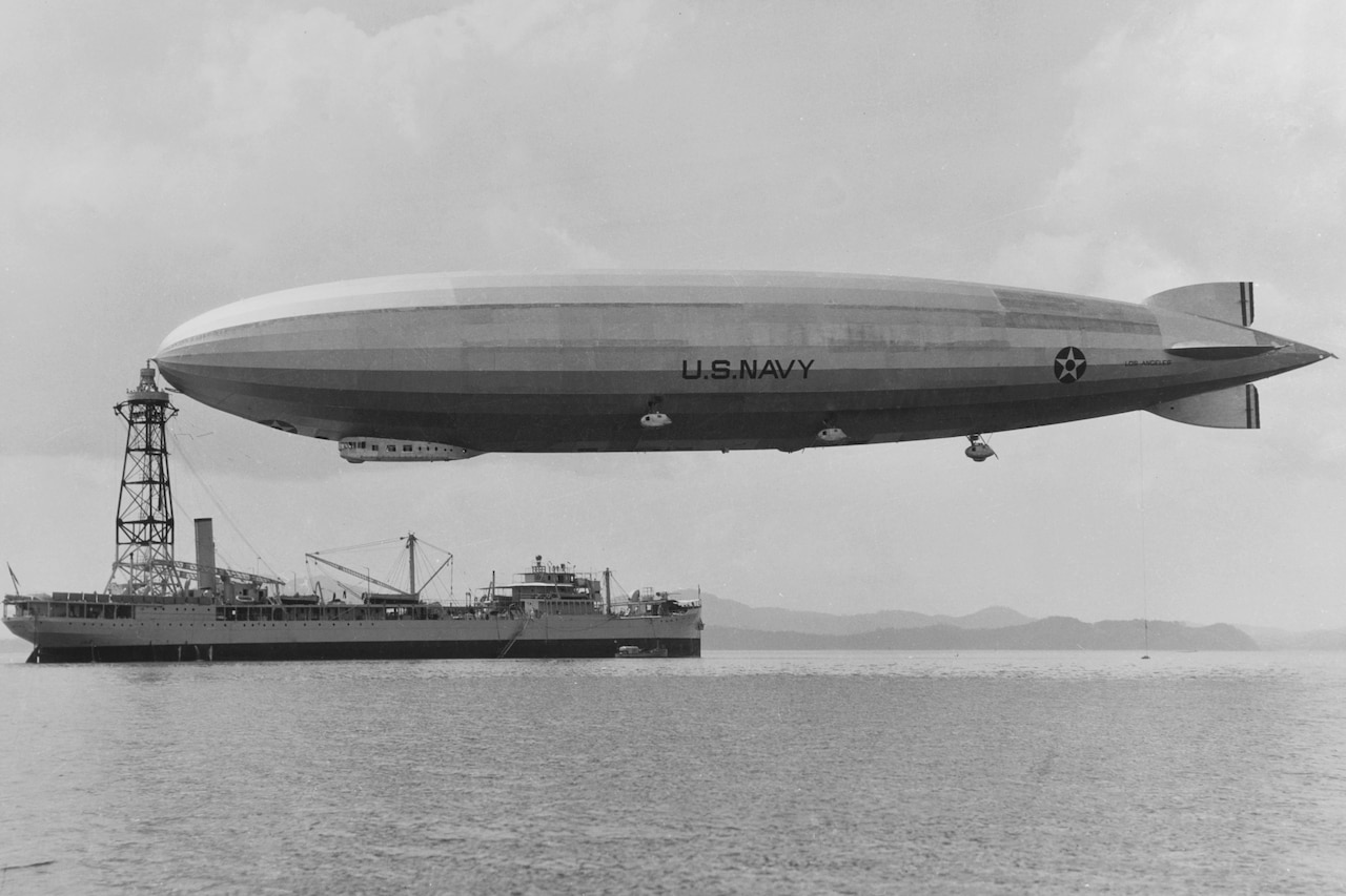 A gigantic airship floats above a military ship in the ocean with the coastline visible in the distance.