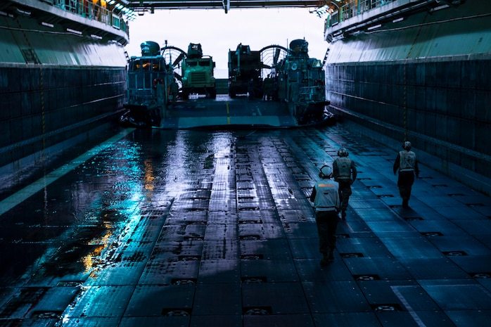 Three people in a dark well deck wearing flight gear and headsets walk toward a landing craft transporting trucks.