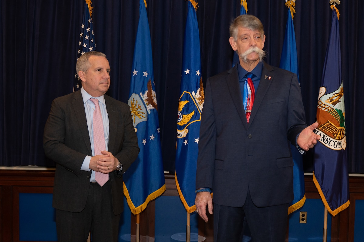 Image of two people speaking in front of flags.