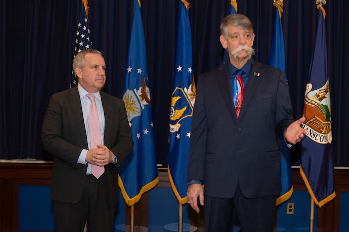 Image of two people speaking in front of flags.
