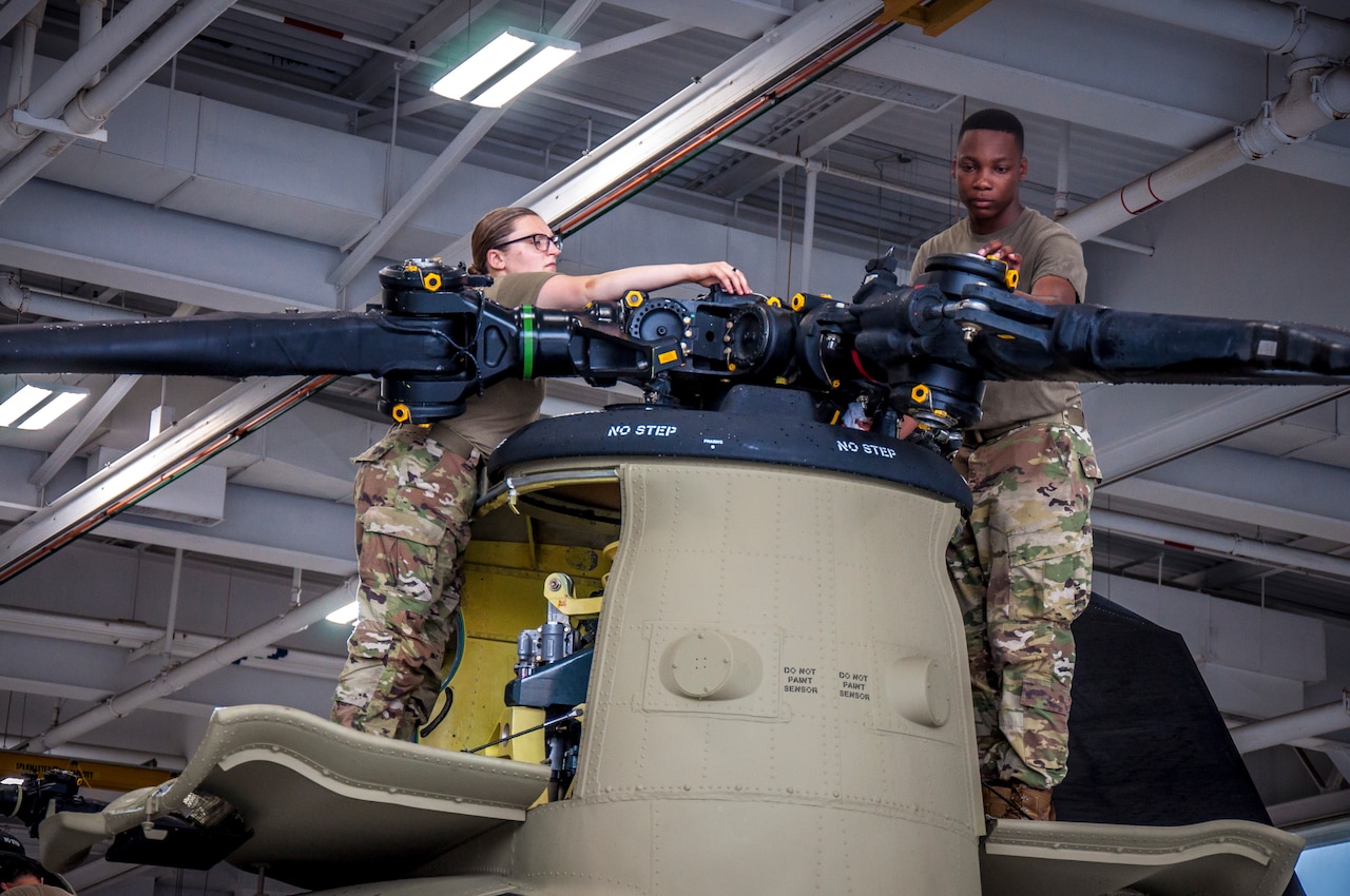 A man and a woman in camouflage military uniforms stand on a helicopter as they repair the rotor blade.