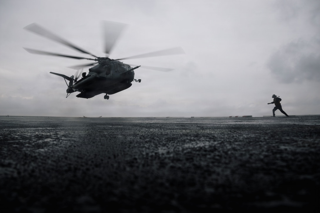 A sailor runs toward a helicopter as it prepares to land on a ship as seen in silhouette.