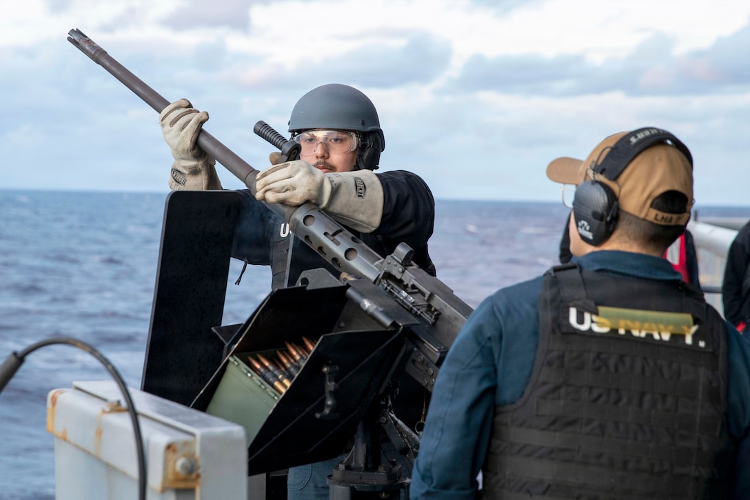 A sailor in protective gear adjusts the barrel of a machine gun as another sailor observes from the side aboard a ship at sea.