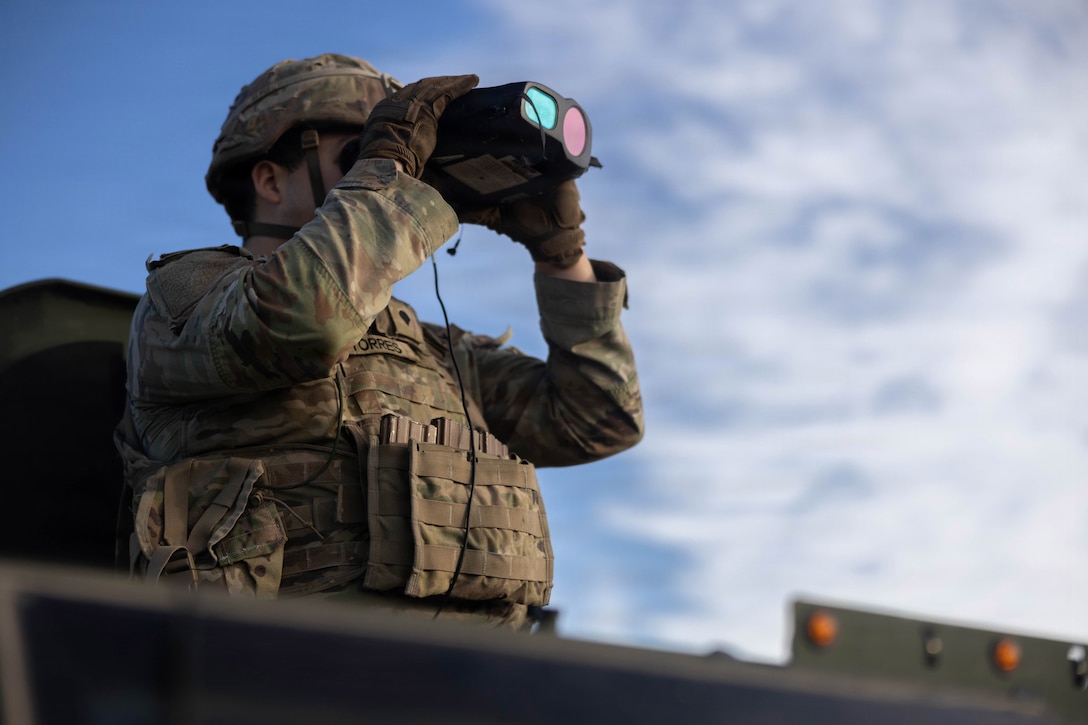 A soldier in camouflage uniform and helmet looks through binoculars while standing near a border fence during the day.