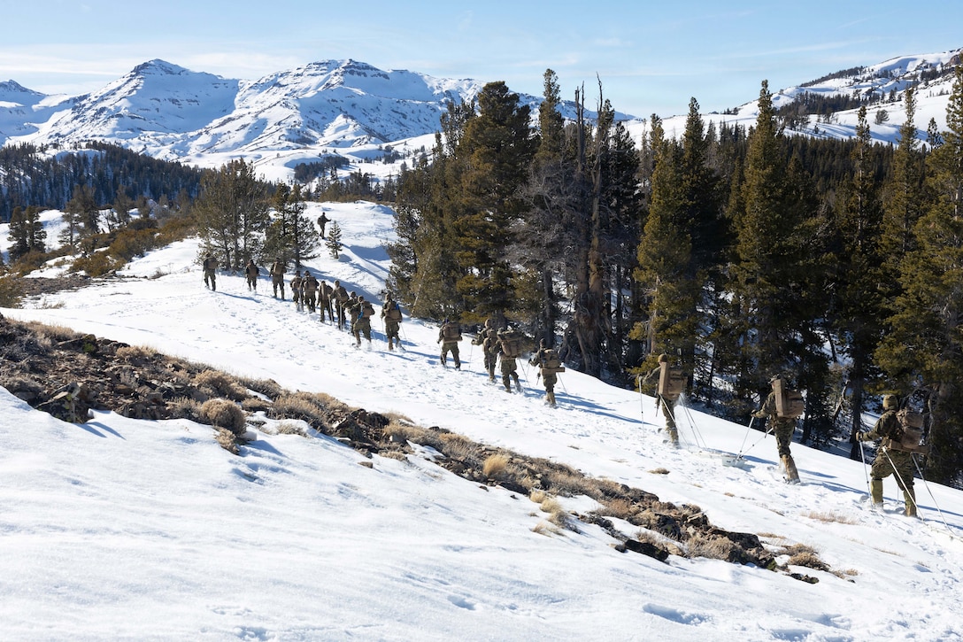 More than a dozen Marines carrying gear hike a snowy mountain under a blue sky.