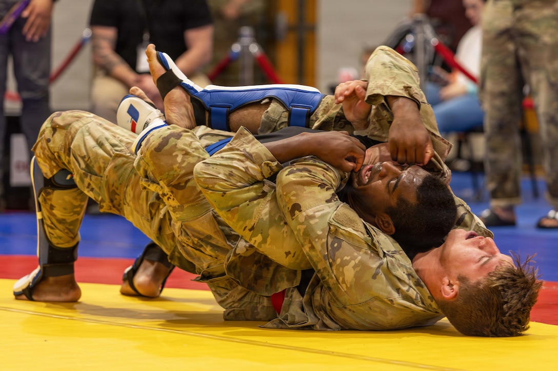 Two soldiers in supine positions grapple on a padded yellow, red and blue mat indoors, with one soldier wrapping an arm around the other's neck as people in the background watch.