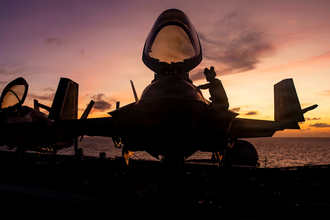 The silhouette of a Marine climbing toward the open cockpit of a fighter jet parked on the deck of a ship is seen, with another jet, water and an orange and purple sky in the background.
