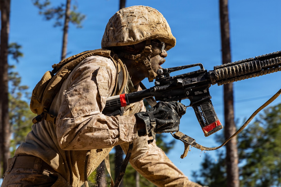 A Marine Corps recruit wearing combat gear splotched with sand holds a rifle with one hand and advances in a wooded area on a sunny day.