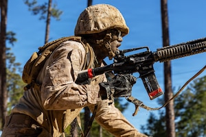 A Marine Corps recruit wearing combat gear splotched with sand holds a rifle with one hand and advances in a wooded area on a sunny day.