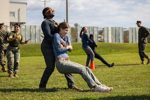 A woman in civilian clothing drags another woman dressed in jeans and a T-shirt, while holding her under the arms and from behind. They are both laughing. Other people, dressed in military uniforms and civilian clothing, mill around in the background.
