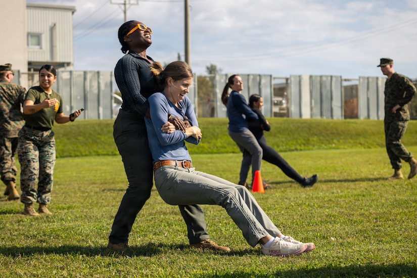 A woman in civilian clothing drags another woman dressed in jeans and a T-shirt, while holding her under the arms and from behind. They are both laughing. Other people, dressed in military uniforms and civilian clothing, mill around in the background.