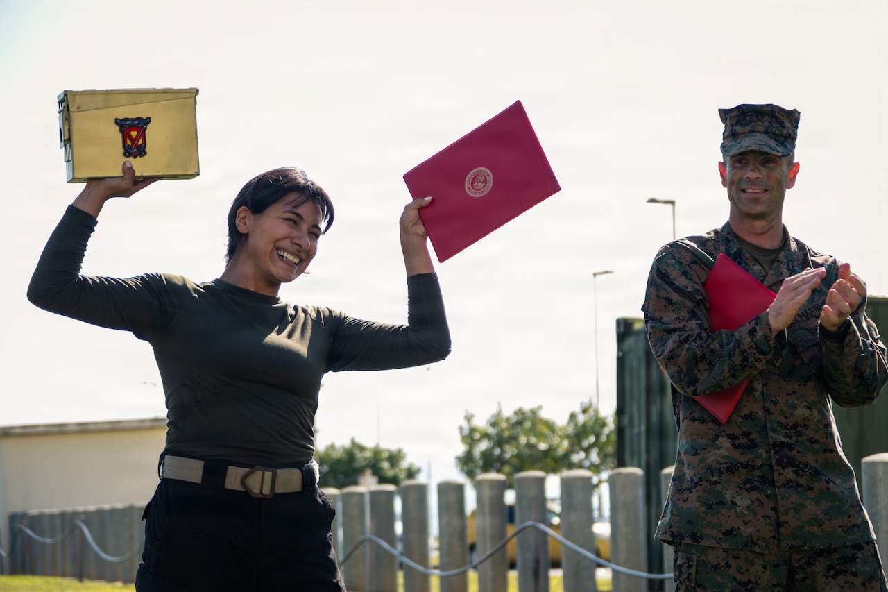 A woman dressed in dark clothing smiles as she holds up a certificate in one hand and a yellow box in another. A man wearing a camouflage military uniform stands next to her and claps.