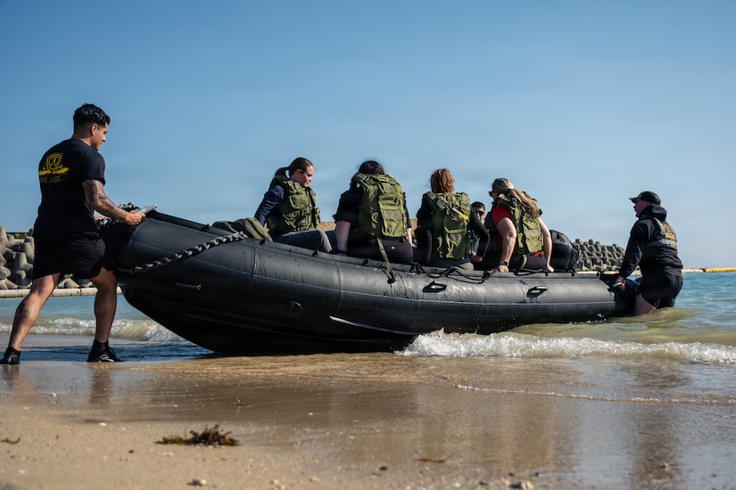 Two men dressed in military physical fitness clothing push a rubber boat into a body of water. People with military vests on are sitting in the boat.