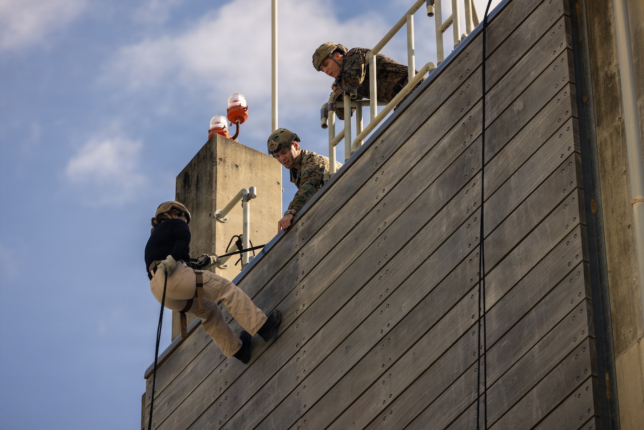 A woman dressed in civilian hiking gear rappels down a wooden wall as two men in military uniforms look down at her.