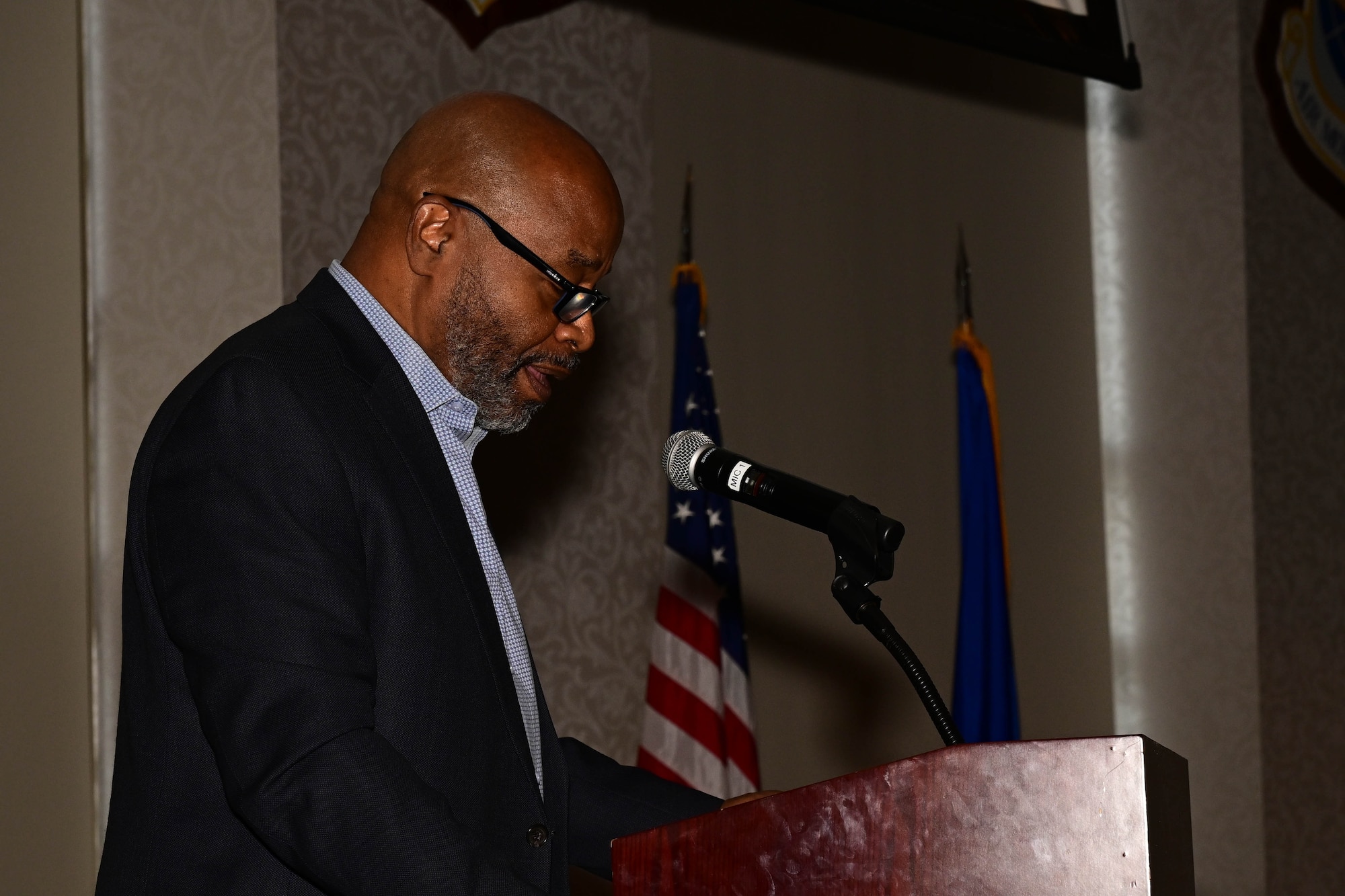 Alonzo Byrd, dressed in a blue dress shirt and dark colored jacket, looking down as he speaks to an audience.