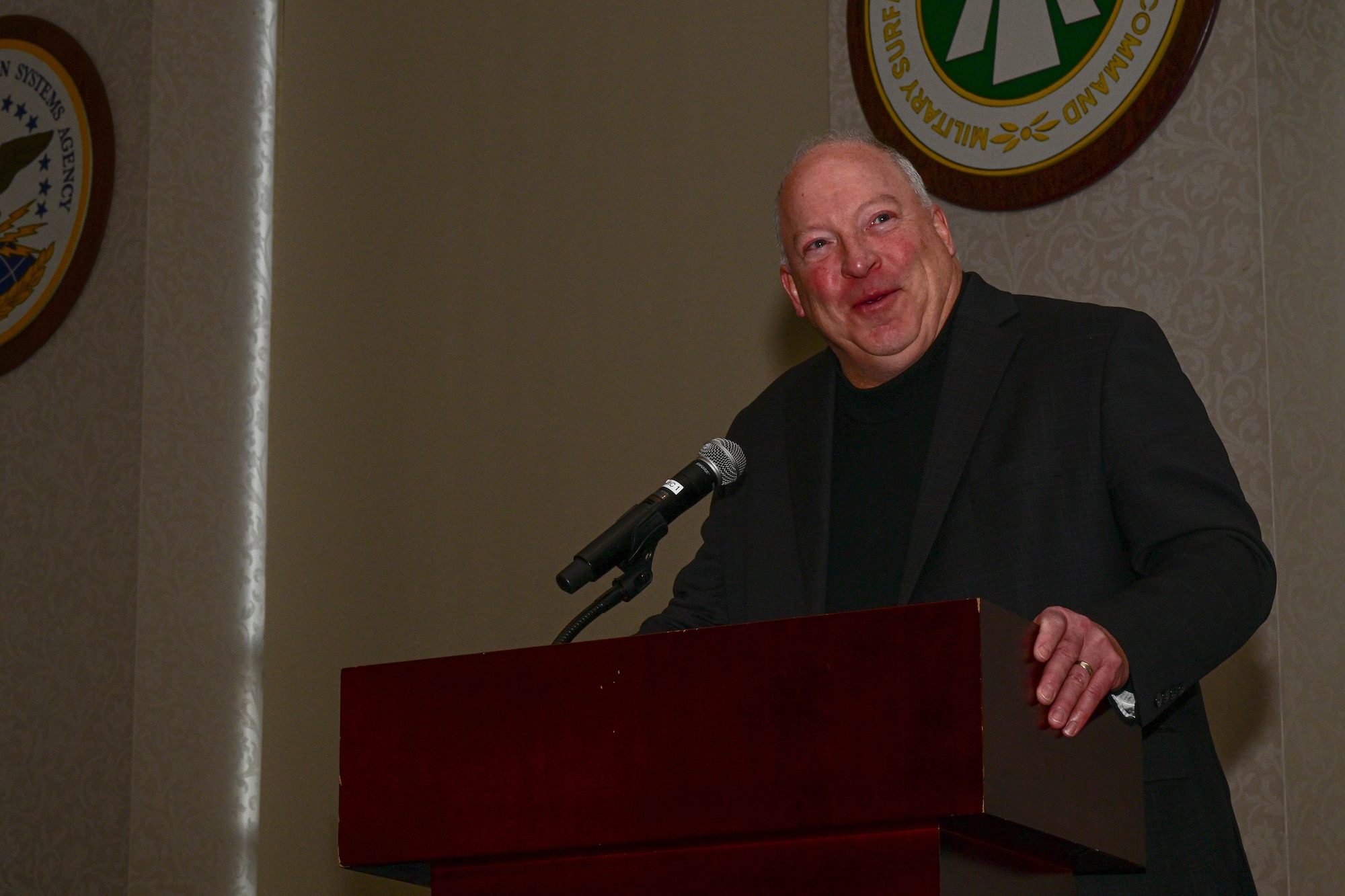 Philip Bissonette, dressed in an all black suit, is smiling while giving a speech at a podium.