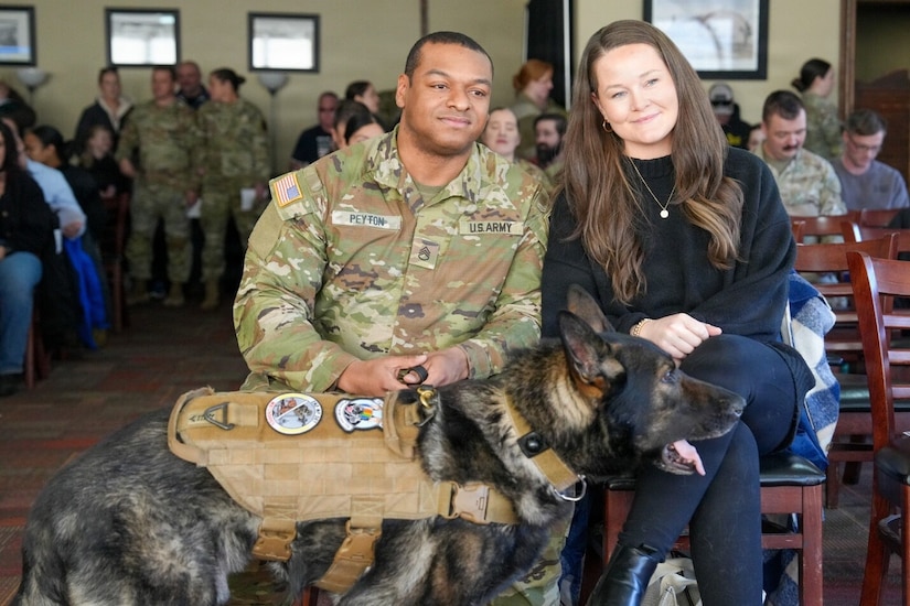 A man dressed in a camouflage military uniform and a woman wearing casual winter attire sit in chairs in a large room. The man is holding a leash attached to a German shepherd that is standing in front of them. There are dozens of people in similar attire sitting and standing behind them.