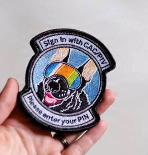 A person holds a colorful patch of a dog wearing protective goggles. The top of the patch reads "Sign in with CAC/PIV," while the bottom reads "Please enter your PIN."