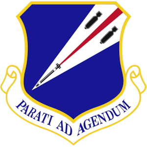 Emblem of the Missouri Air Natioanl Guard's 131st Bomb Wing. The emblem is shaped like a shield. On it, in a royal blue field, three black bombs fall from top right to bottom left leaving red and white streaks. A scroll underneath the shield reads "Parati ad Agendum" in royal blue.
