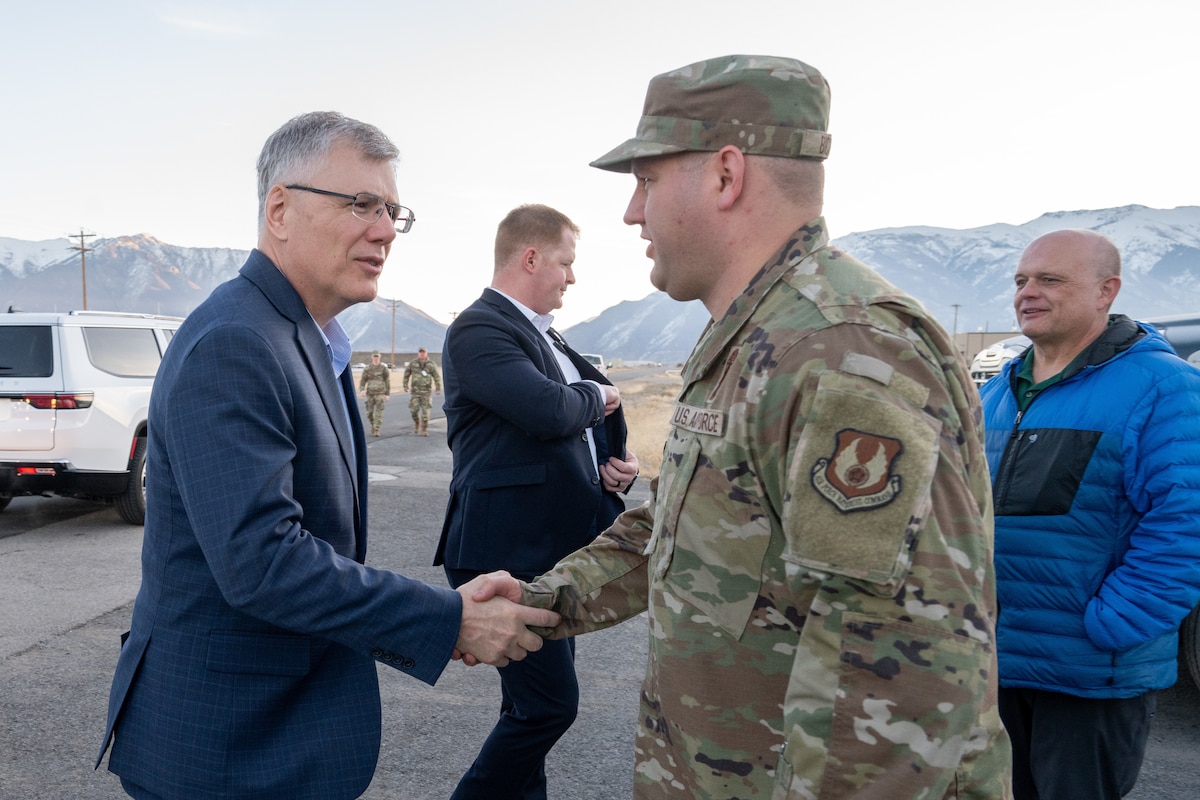 A man in a business suit shakes hands with a uniformed Airman outdoors with mountains in the background