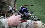 Soldier in camouflage holding a small quadcopter drone with visible wiring and a strapped-on battery outdoors.