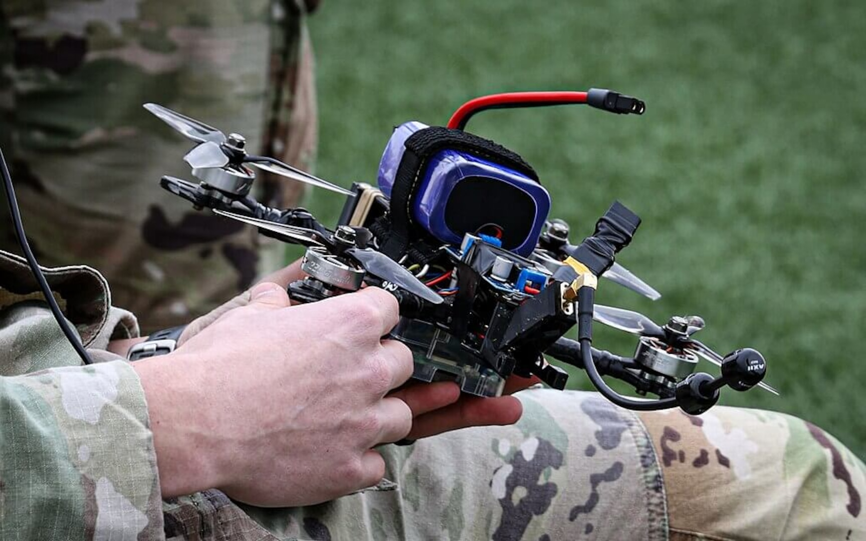 Soldier in camouflage holding a small quadcopter drone with visible wiring and a strapped-on battery outdoors.