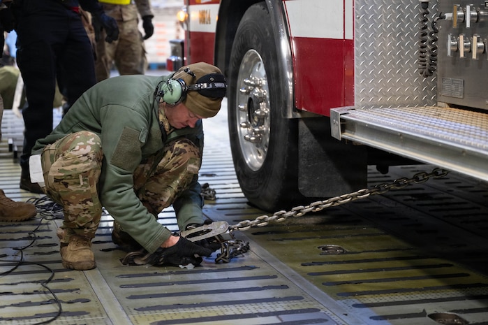 An Airman crouches on the ground next to the wheel of a firetruck, attaching a chain to the floor of a C-17 aircraft to secure the vehicle.