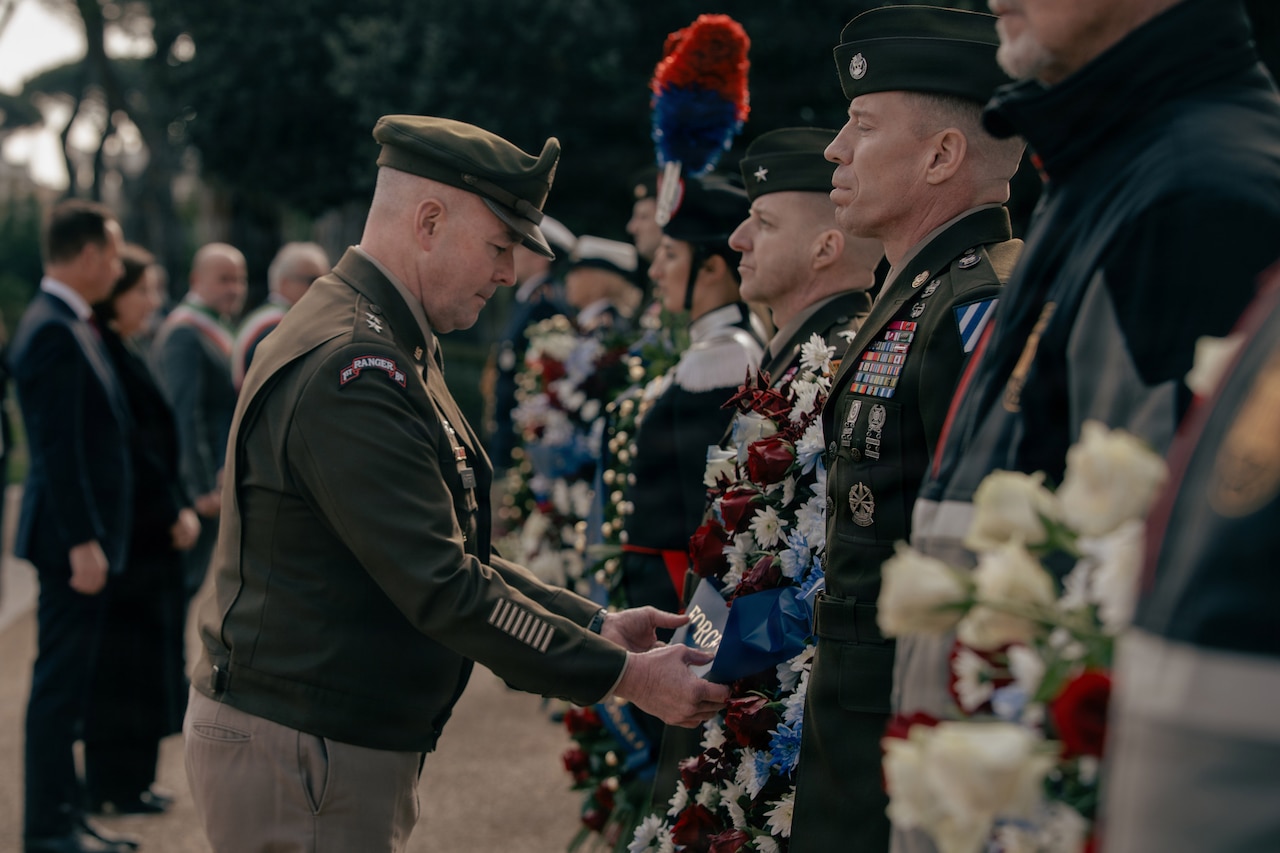 A man in a military dress uniform inspects a wreath during a ceremony at a monument in Italy. There eight people in military dress uniforms standing in a line with wreaths in between them. There are four other people in business attire standing in the background.