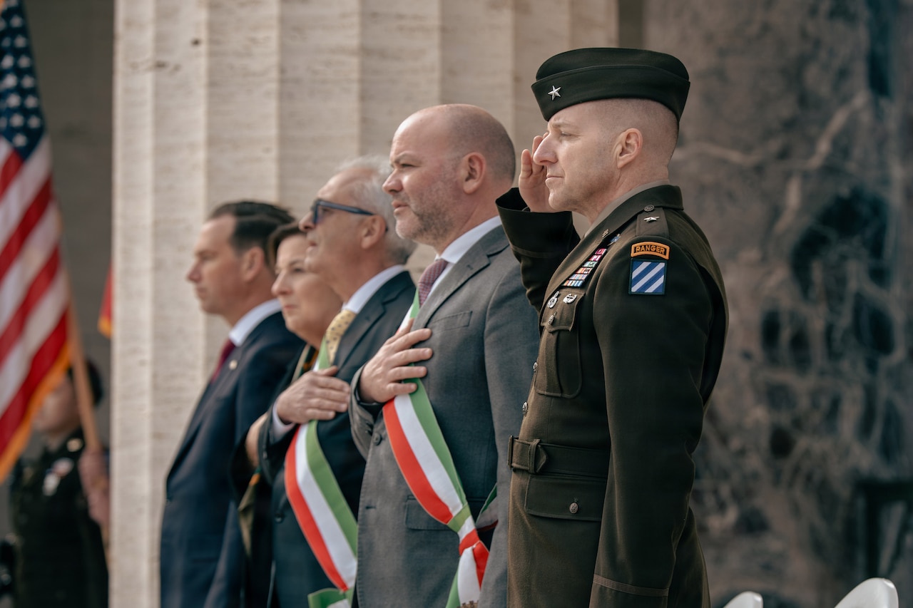 A man wearing a military dress uniform renders a salute while two men in business attire stand with hands over their hearts during an outdoor commemoration ceremony in Italy. Another man and a woman in business attire stand beside them in a row. The American flag is displayed in the background.