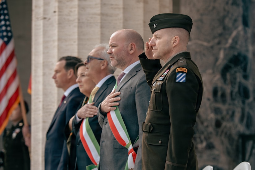 A man wearing a military dress uniform renders a salute while two men in business attire stand with hands over their hearts during an outdoor commemoration ceremony in Italy. Another man and a woman in business attire stand beside them in a row. The American flag is displayed in the background.