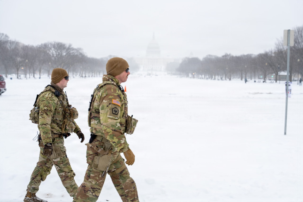 Two people in camouflage military uniforms walk along a snow-covered sidewalk in front of the U.S. Capitol.