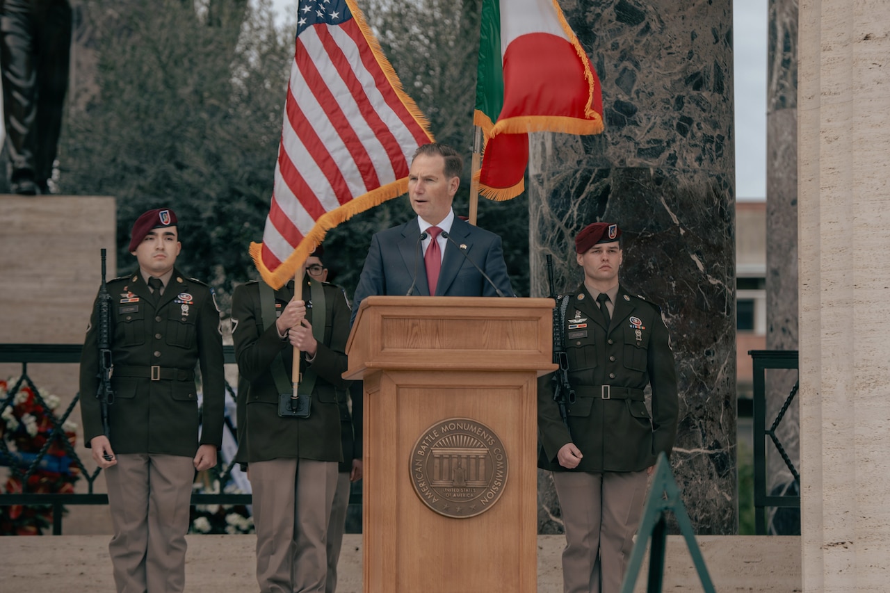A man in a suit speaks behind a lectern as four people in military dress uniforms stand behind him. One of them in holding the American flag while another holds the Italian flag.